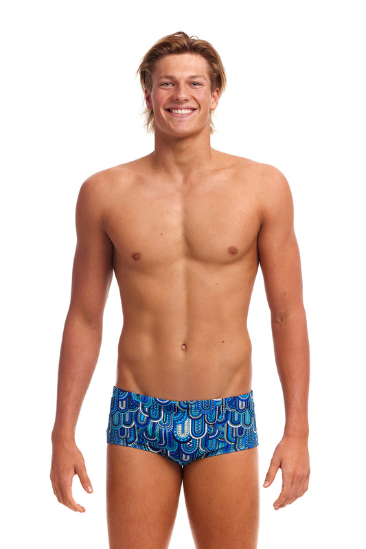 A young man with blond hair smiles at the camera, wearing Funky Trunks Men's Flight School Classic Trunk known for their chlorine resistance. He stands against a white background, exuding a casual, confident demeanor.