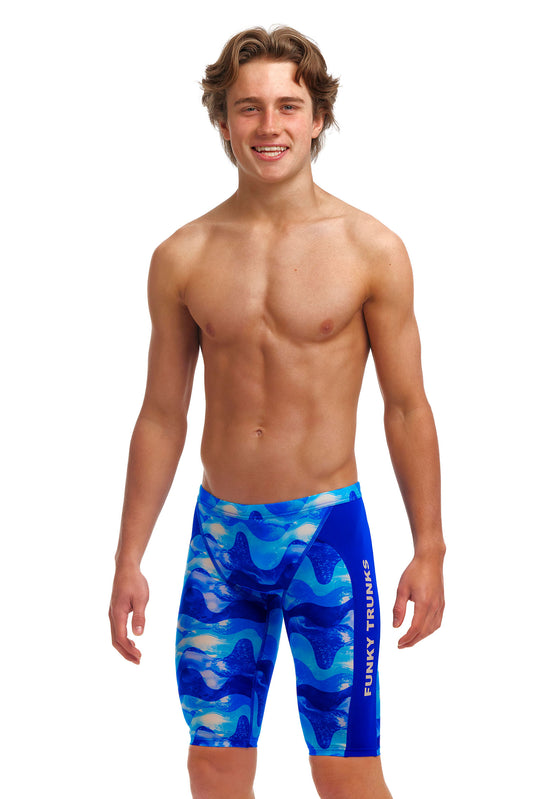 A young man wearing blue patterned Funky Trunks Boys Dive In Training Jammer swim trunks smiles while posing against a white background.