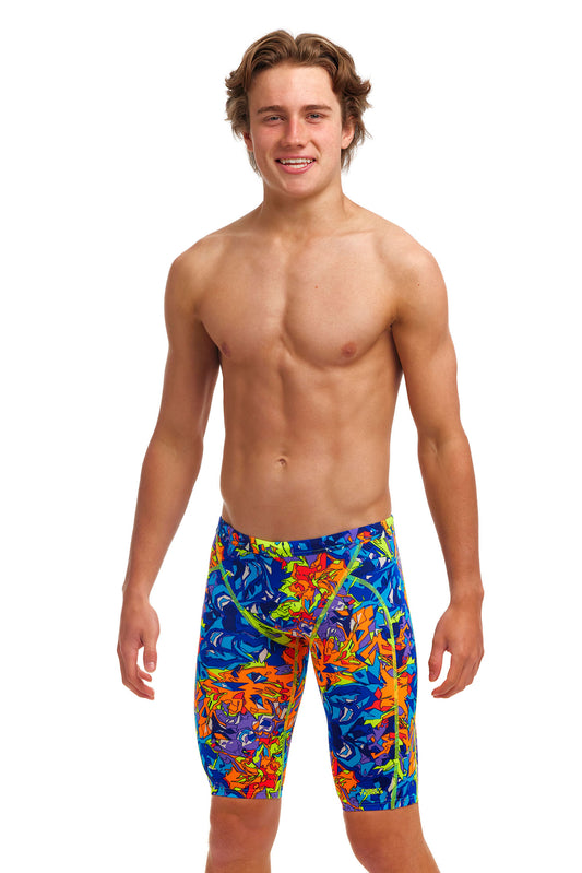 A young man with tousled hair smiling at the camera, wearing colorful knee-length board shorts made from Funky Trunks ECO C-INFINITY fabric for excellent chlorine resistance, standing against a white background.