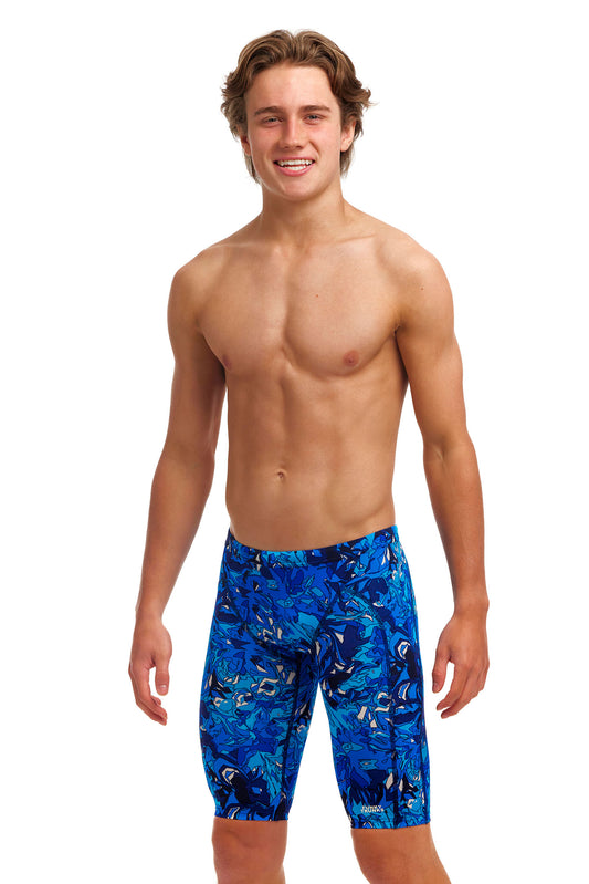 A young man with long curly hair, smiling, wearing blue swimming trunks made from ECO C-INFINITY for exceptional chlorine resistance, showcasing a vibrant tropical fish print, standing against a white background.